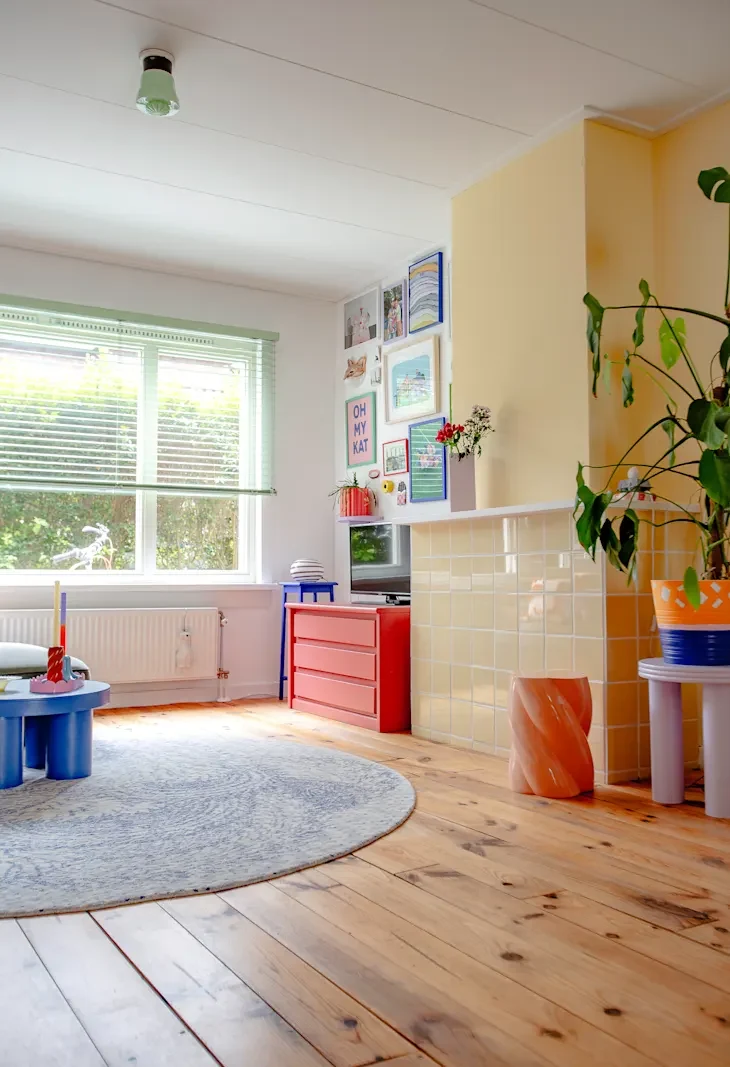 Bright living room featuring a blue coffee table, red dresser, and yellow wall with framed art. A large plant and round rug add warmth.