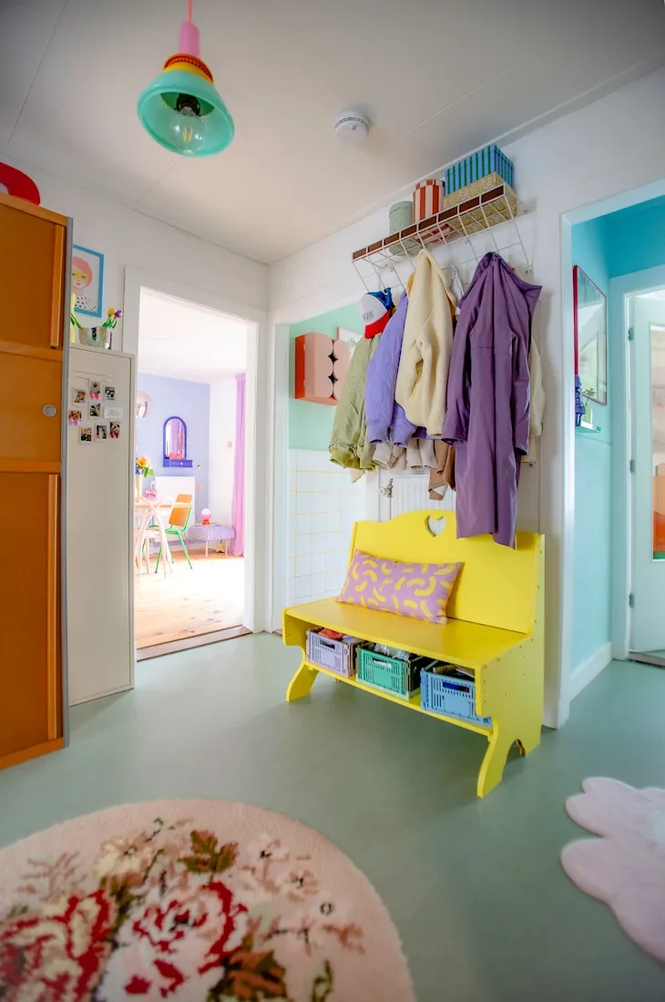 Colorful entryway featuring a yellow bench with a patterned pillow, hanging coats, and a floral rug.