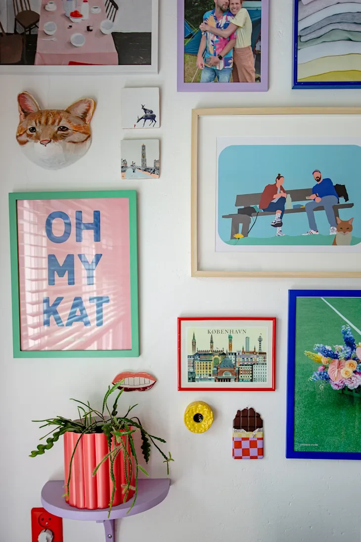 Colorful wall decor featuring framed art, a cat sculpture, and a potted plant on a purple shelf.