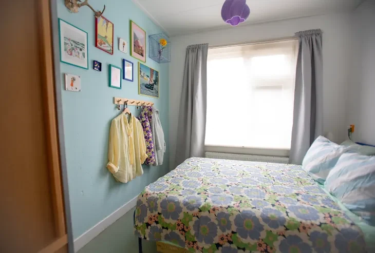 Cozy bedroom with a floral quilt, wall art, hanging clothes, and a purple pendant light.