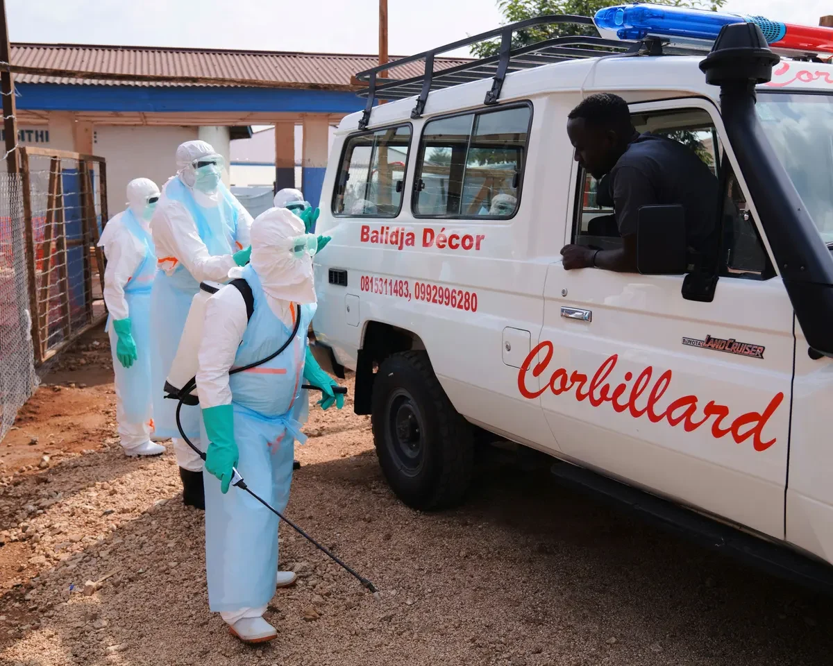 Healthcare workers wearing full protective suits and masks stand beside a white ambulance