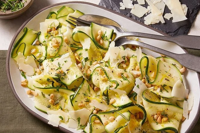 Overhead horizontal image of a white oval serving dish filled with ribbon-cut zucchini salad. The salad is garnished with pine nuts, shaved parmesan cheese, fresh parsley and thyme, and lemon zest, with a fork and spoon resting in the dish.