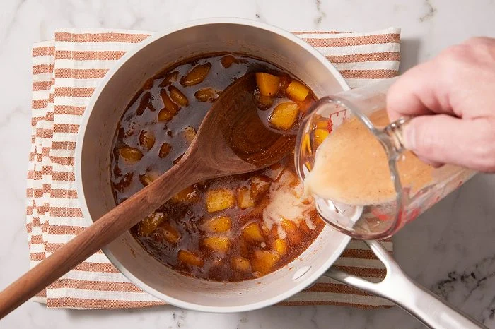 Overhead shot of a fruit filling simmering in a saucepan as a cornstarch slurry is poured in from a measuring cup to thicken the mixture.