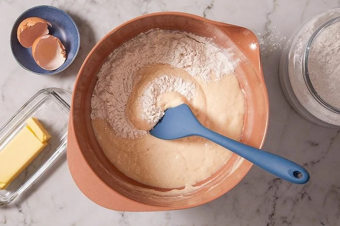 Overhead shot of batter being folded with flour in a large mixing bowl using a blue silicone spatula.
