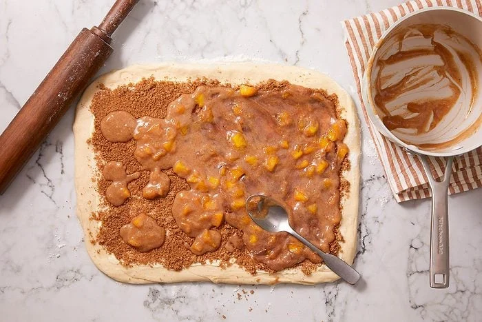 Overhead shot of rolled dough topped with a layer of brown sugar and fruit filling, as the filling is spread evenly with a spoon.