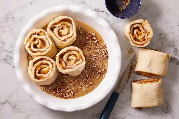 Overhead shot of sliced fruit-filled dough rolls arranged in a buttered baking dish, with chopped pecans sprinkled over the top.