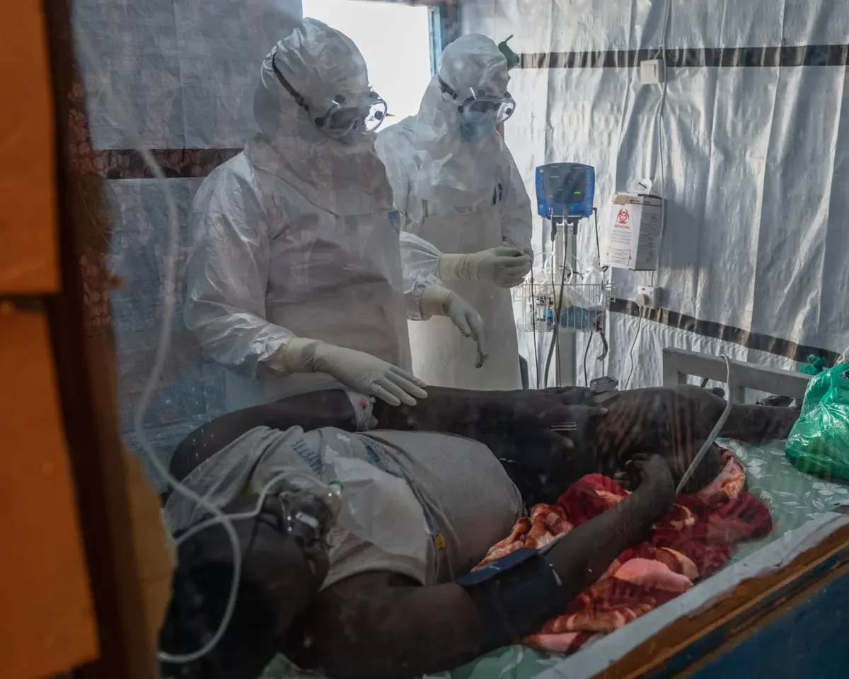 Two healthcare workers in full protective suits stand beside a patient lying on a hospital bed.